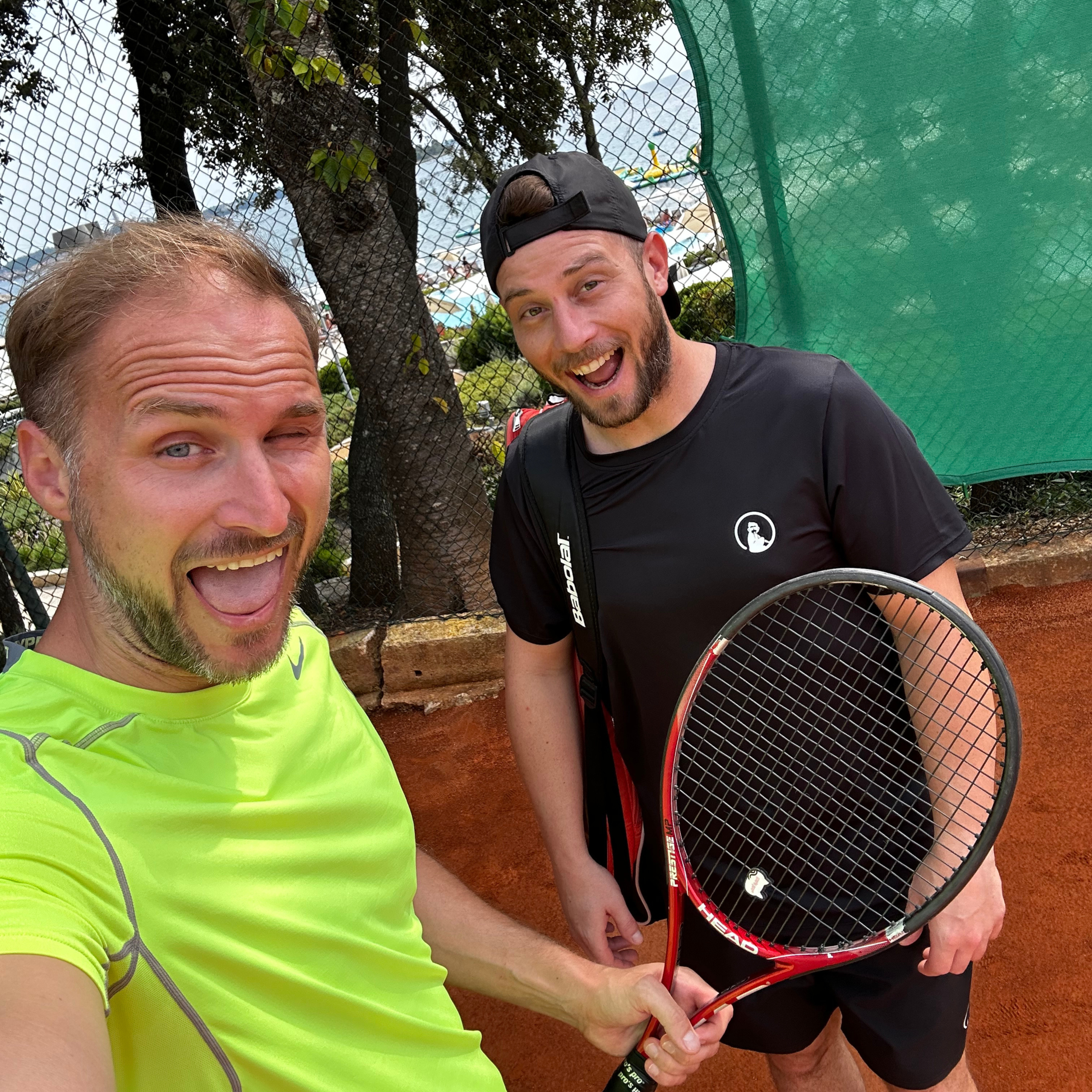 Two colleagues laughing on a clay tennis court holding rackets